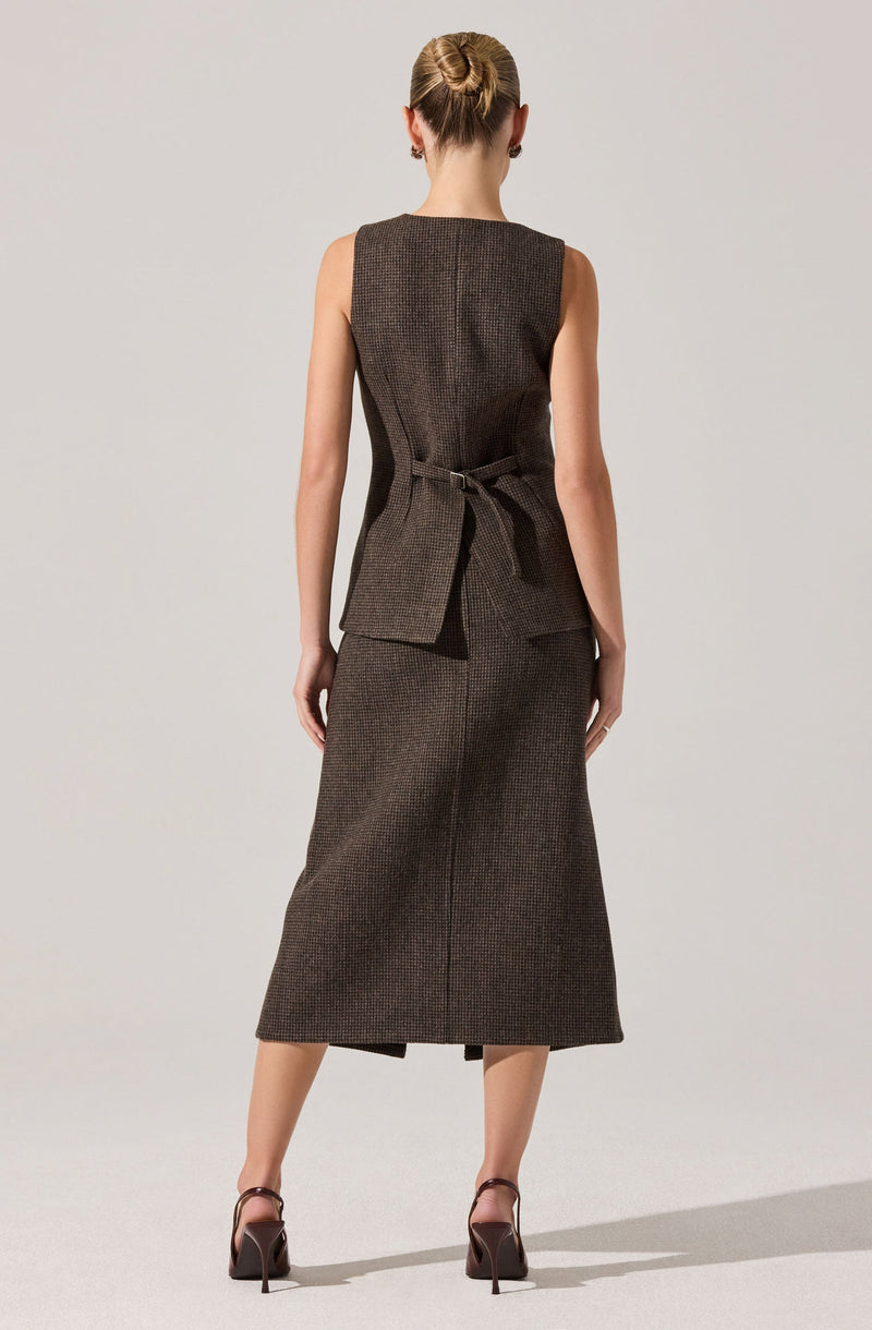The outfit consists of a sleeveless, tailored vest with a high neckline and a belt tied at the waist, matching a midi-length skirt with a subtle textured pattern. The person is wearing dark, strappy high-heeled shoes.