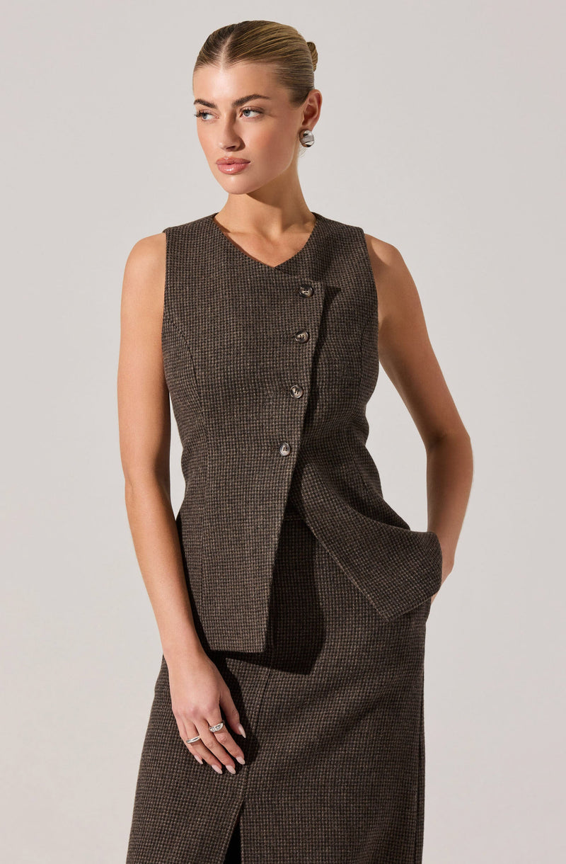 A woman is wearing a sleeveless, knee-length skirted suit in a dark, checkered pattern. The suit has a tailored top with an asymmetrical button closure and a matching skirt with a front slit. She has her left hand in her skirt pocket and is accessorized with earrings and rings.