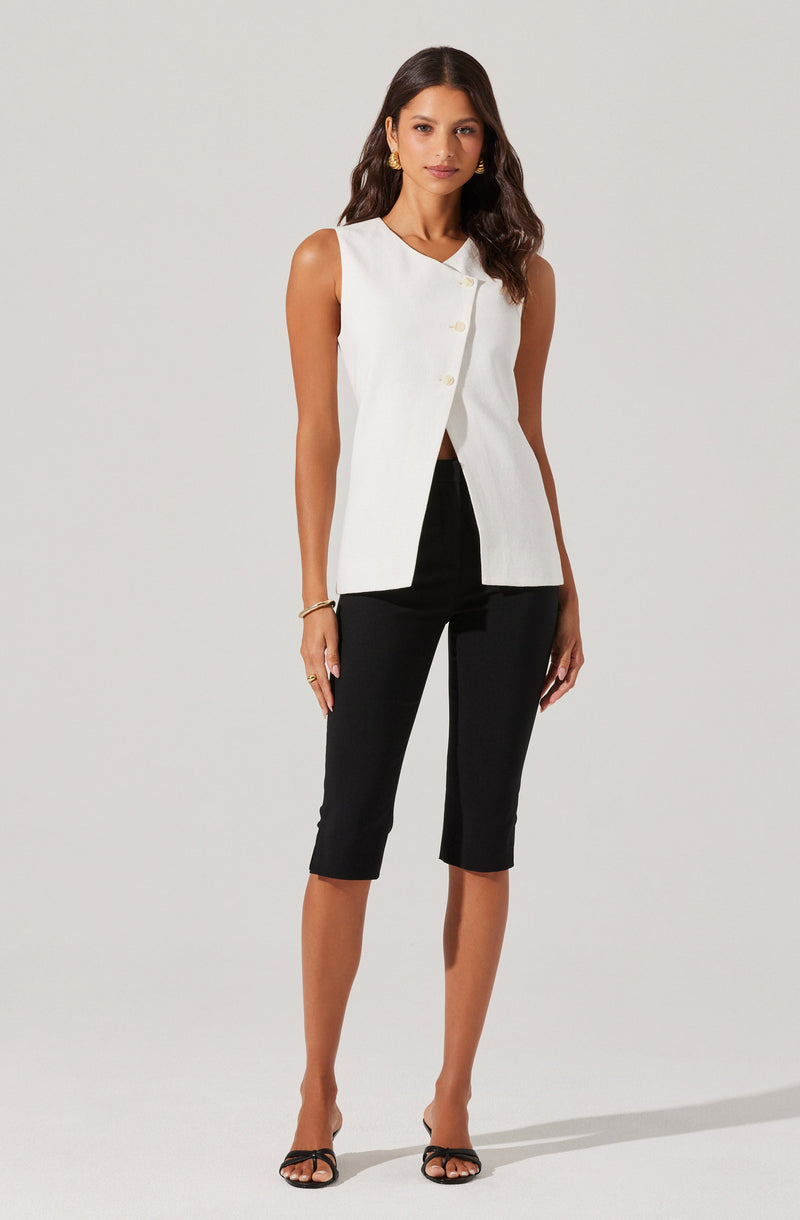 A woman is wearing a sleeveless white vest with a high collar and large buttons down the front, paired with black cropped pants that reach just below the knees. She completes her outfit with open-toe black heels.