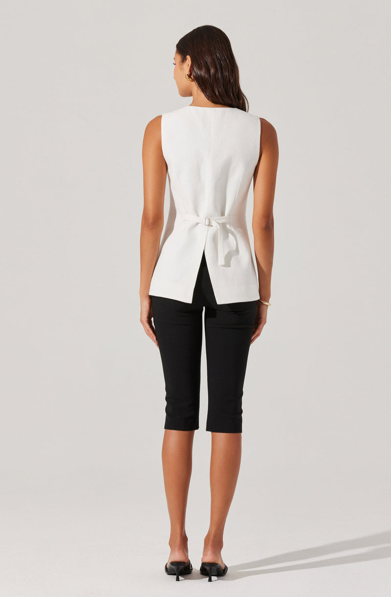 A woman is wearing a sleeveless, white, tailored top with a back tie detail, paired with black, knee-length fitted pants. She has dark, shoulder-length hair and is wearing small earrings and a bracelet on her left wrist. She is standing against a plain, light-colored background in a relaxed pose.