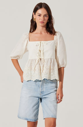 A woman is wearing an off-white, square-neck blouse with puffed three-quarter sleeves. The blouse features decorative eyelet embroidery and a ruffled hem, with ties at the front for adjustment. She is pairing it with light-wash denim shorts.
