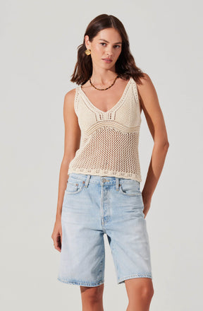 A woman wearing a sleeveless, cream-colored, crocheted tank top with a V-neckline and intricate pattern design, paired with light-wash denim Bermuda shorts. She accessorizes with gold earrings and a beaded necklace.