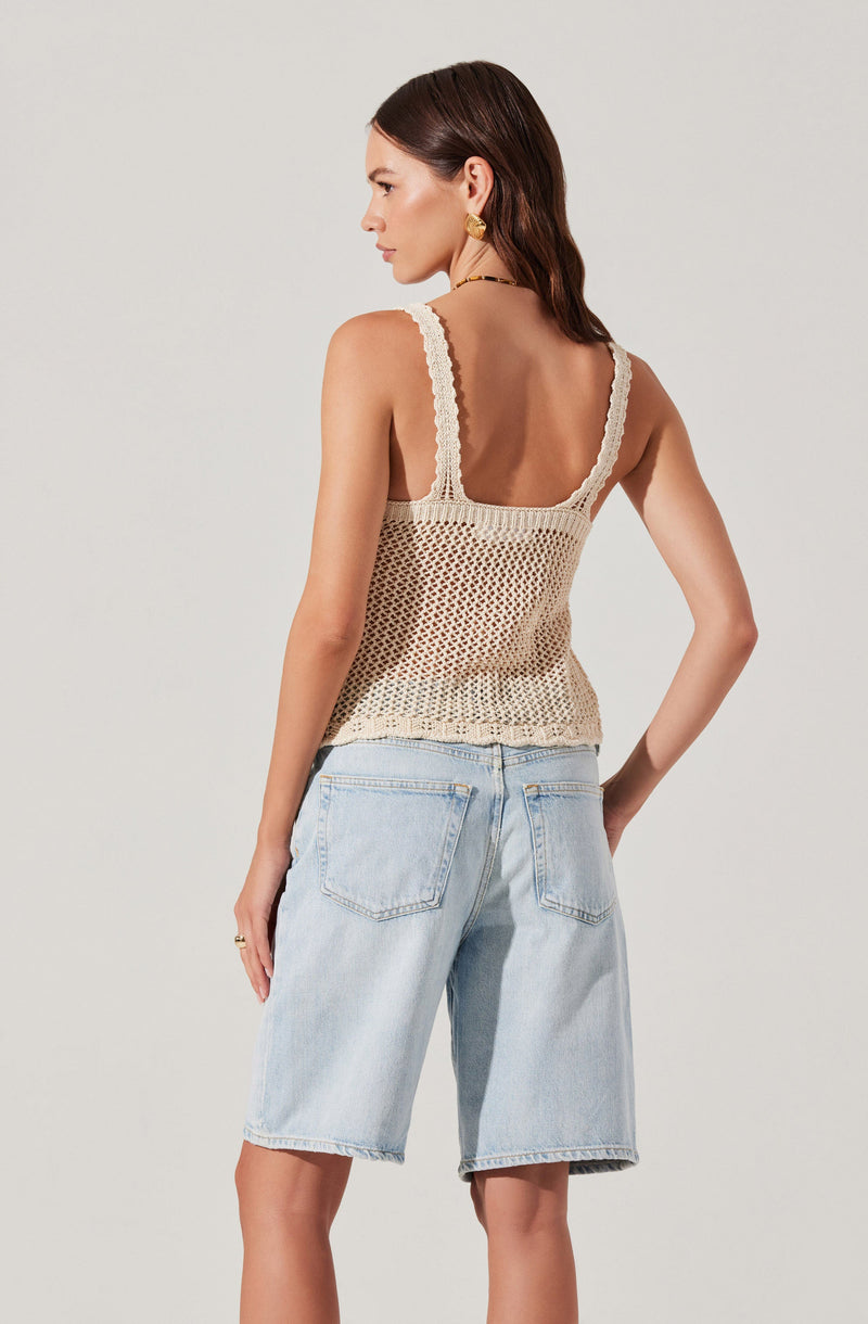 A woman is wearing a sleeveless, beige, crocheted top with wide straps and a square neckline. She pairs the top with light blue denim shorts that have back pockets. Her hair is styled in loose waves, and she is accessorized with gold earrings.
