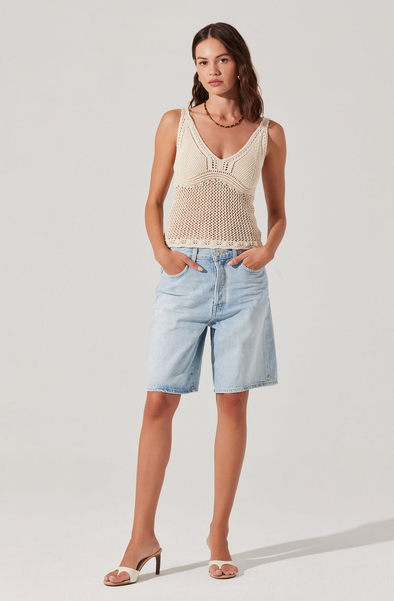 A woman is wearing a sleeveless, cream-colored, crocheted knit top with a V-neckline and intricate open-knit patterns. She has paired it with light-wash, knee-length denim shorts and nude open-toe high-heeled sandals.