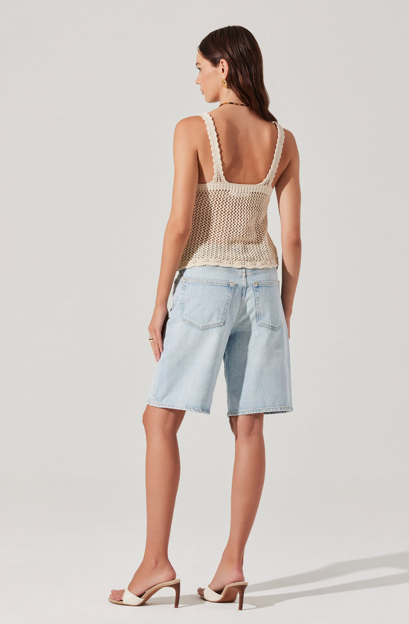 A woman is wearing a sleeveless, cream-colored crochet top with thick straps and a loose fit. She pairs it with light-wash denim shorts that have a relaxed fit and large back pockets. She completes her outfit with white, open-toed high-heeled sandals featuring a single strap across the toes and a slim heel.