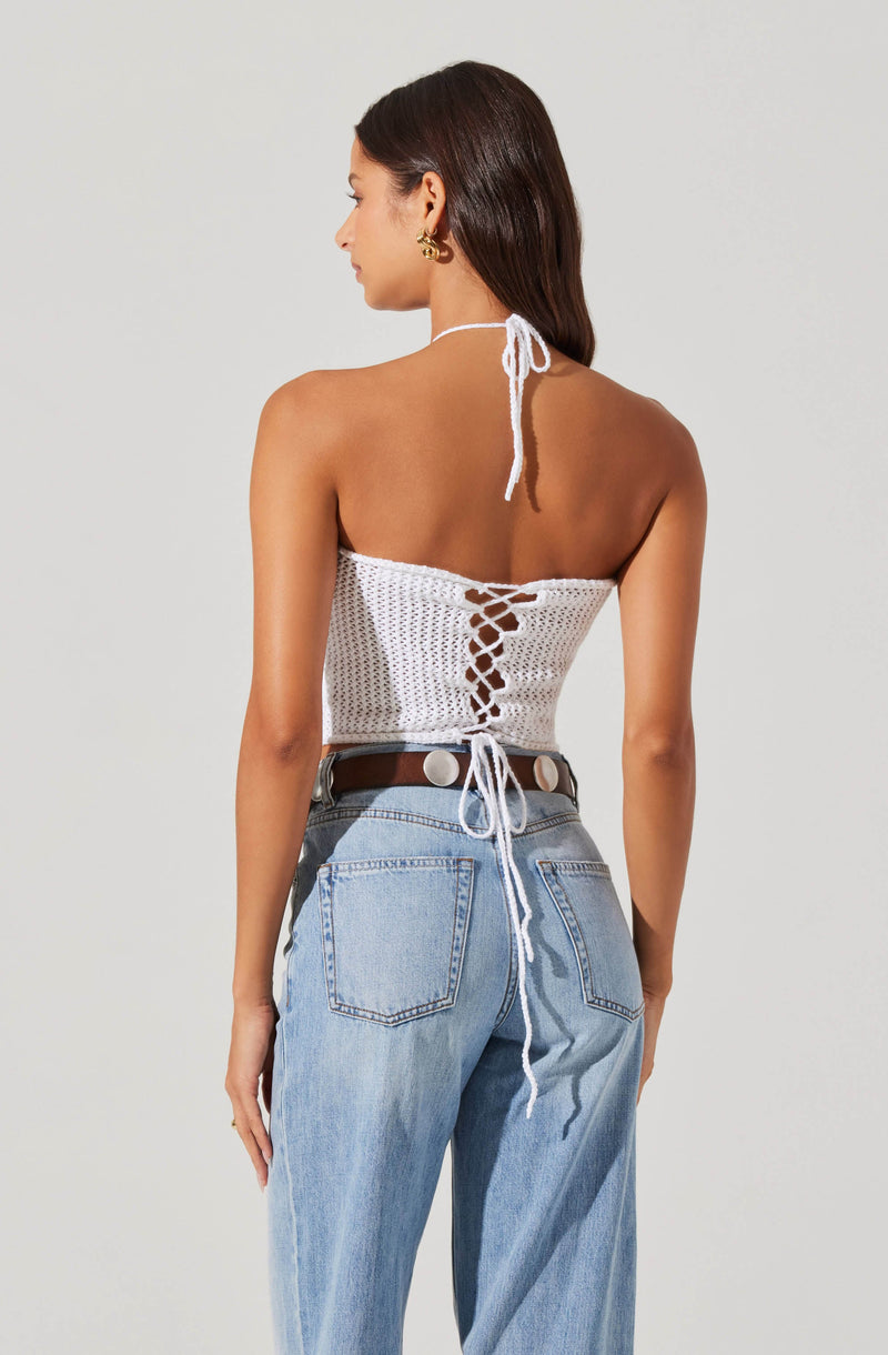 A woman is wearing a white crocheted halter top with a lace-up back detail tied at the neck and waist. She pairs the top with light blue high-waisted jeans and a dark brown belt with large circular silver belt loops.
