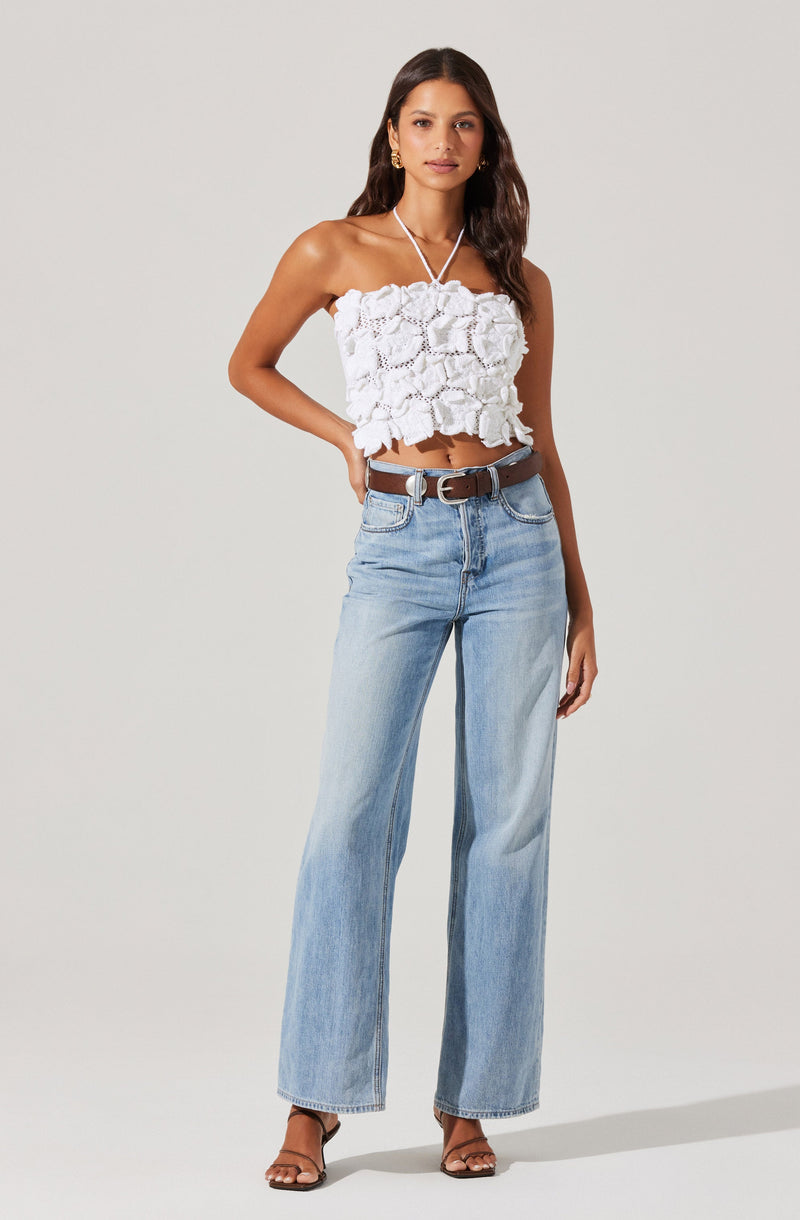 A woman is wearing a white, strapless, ruffled crop top with a thin halter strap around her neck. She has paired it with light blue, high-waisted, wide-leg jeans and a brown belt. She is also wearing black strappy high-heeled sandals and gold hoop earrings.
