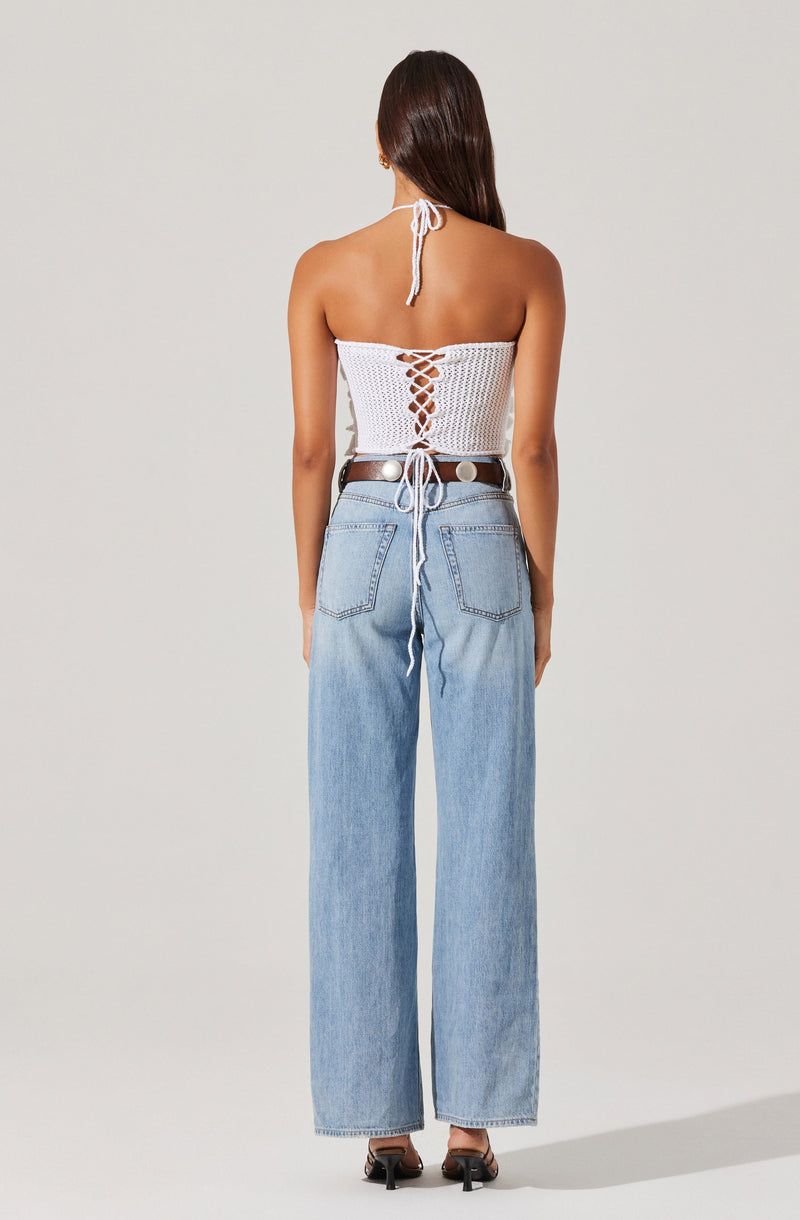 A woman is wearing a white, halter-style top with a open back and crisscross lace-up detail, paired with wide-leg, light blue denim jeans featuring back pockets. She is standing barefoot against a plain light-colored background.