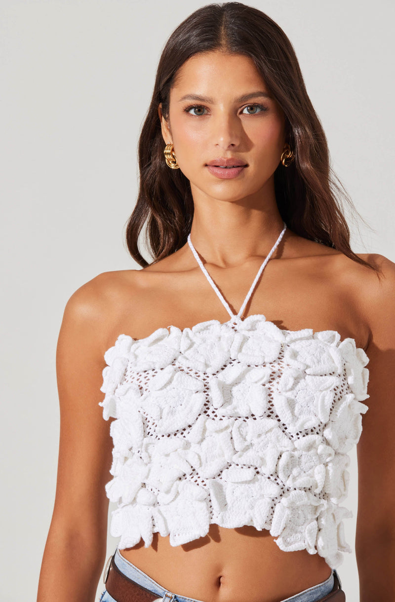 A woman is wearing a sleeveless, strapless white top made of textured fabric with large, ruffled floral appliqués and open-knit details. She accessorizes with gold hoop earrings and a white, knitted tie around her neck.