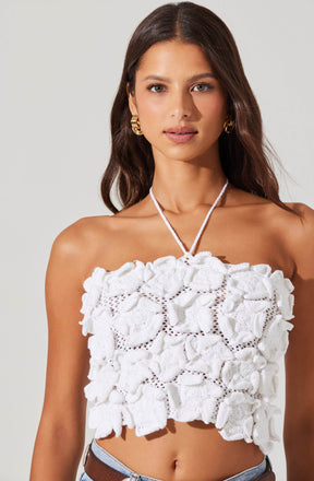 A woman is wearing a sleeveless, strapless white top made of textured fabric with large, ruffled floral appliqués and open-knit details. She accessorizes with gold hoop earrings and a white, knitted tie around her neck.