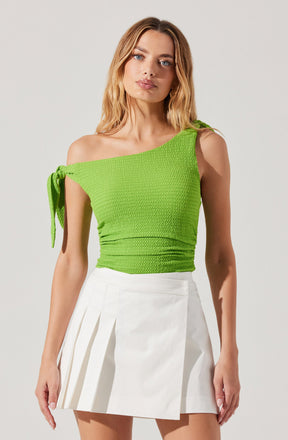 A woman is wearing a bright green, textured, sleeveless top with a one-shoulder design. The top features a tied shoulder strap on her left side. She pairs it with a high-waisted, white mini skirt that has pleats on the left side.