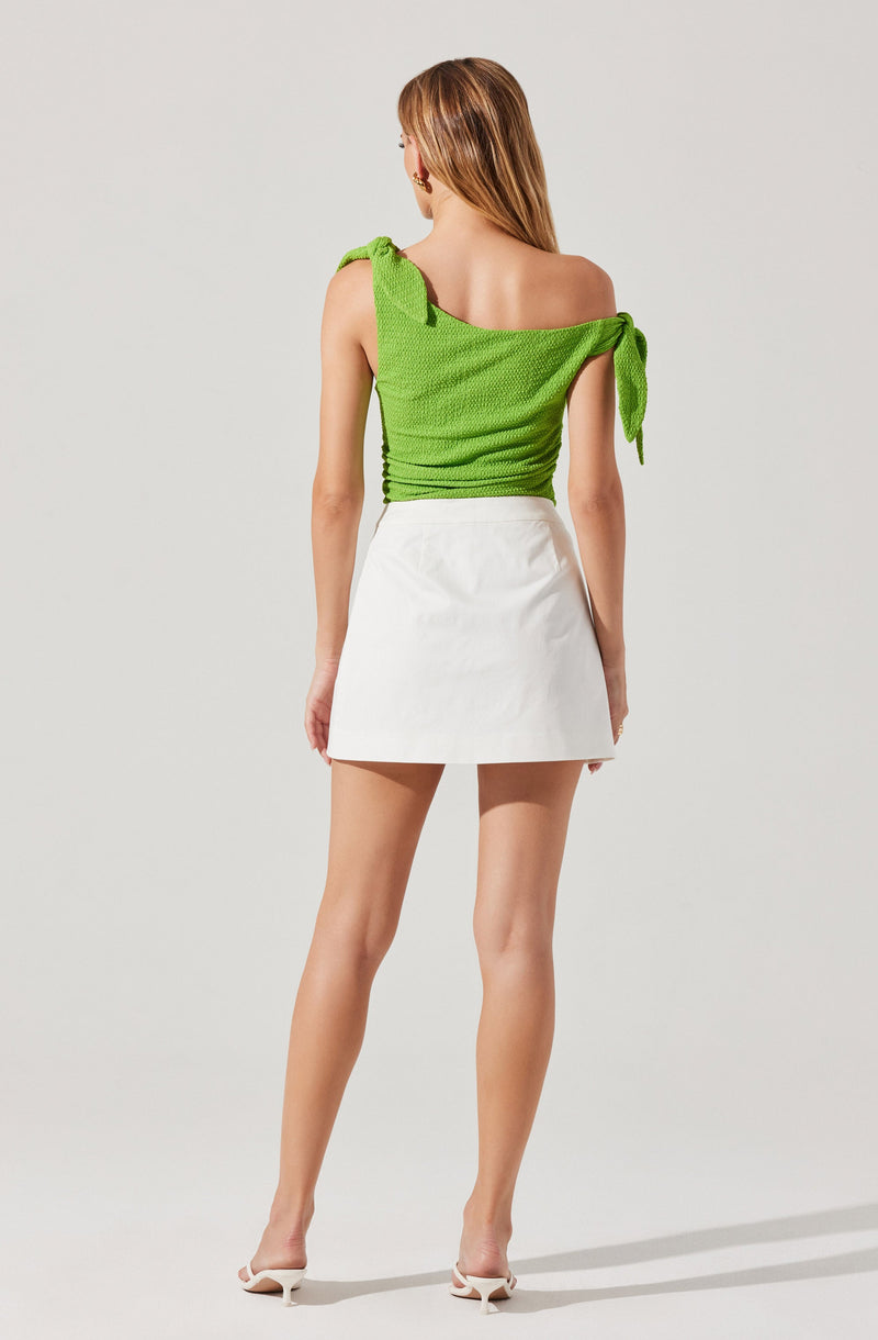 The image shows a woman dressed in a bright green, textured top with ruffled, tied shoulder straps, paired with a white, high-waisted mini skirt. She is wearing white high-heeled shoes and has long, straight hair.