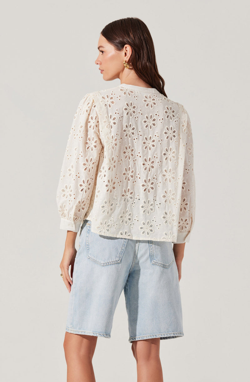 A woman is wearing a white, long-sleeved embroidered blouse featuring a floral cut-out pattern. The blouse has puffed sleeves with elastic cuffs and a relaxed fit. She pairs it with light-wash denim shorts that have a classic five-pocket design and a slightly distressed look. She is accessorized with gold hoop earrings.