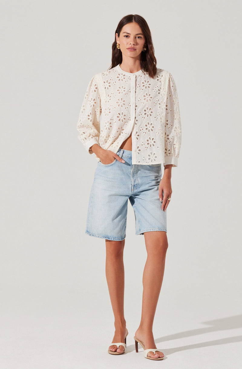 A woman is wearing a white, long-sleeved, button-up blouse with eyelet lace pattern and floral cutouts. She has paired the blouse with light-wash denim shorts and nude high-heeled sandals.