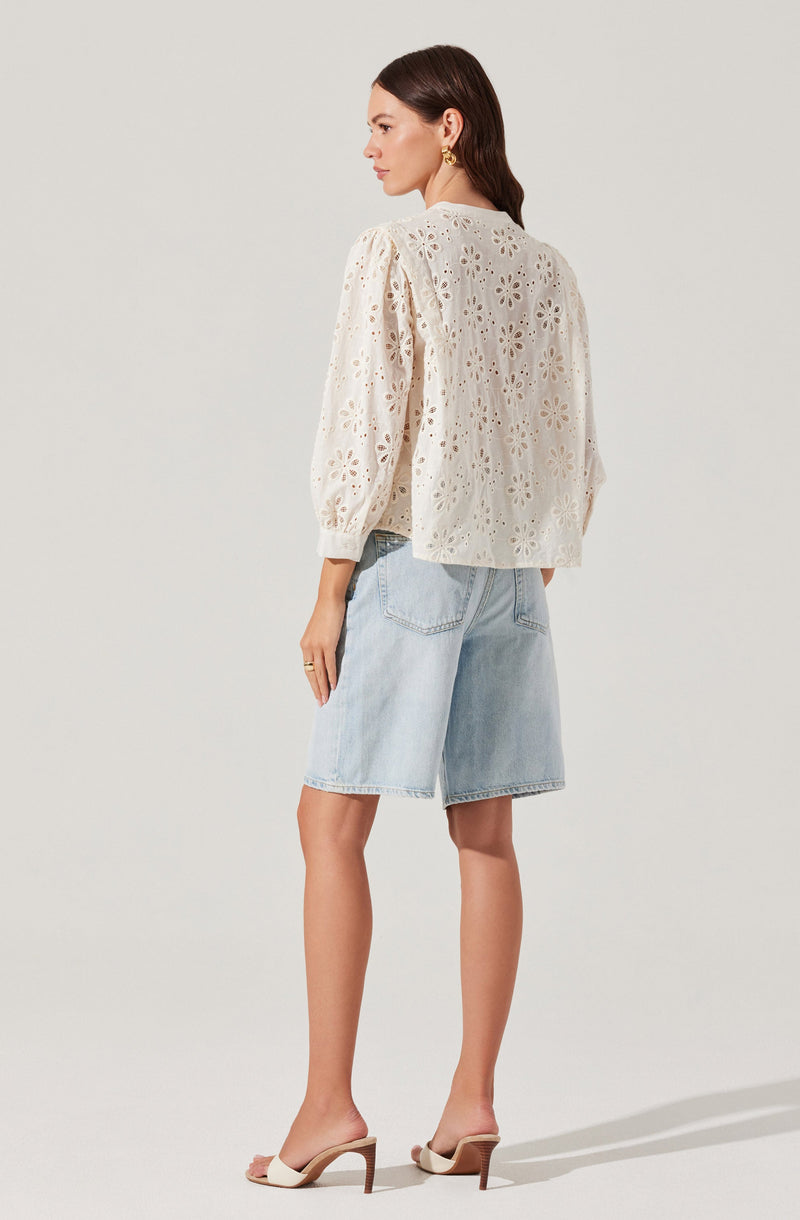 A woman is wearing a cream-colored, long-sleeved blouse with embroidered floral cut-out patterns. The blouse has a loose fit with balloon sleeves gathered at the cuffs. She pairs it with light wash denim Bermuda shorts. She completes the outfit with open-toed high-heeled sandals and gold drop earrings.