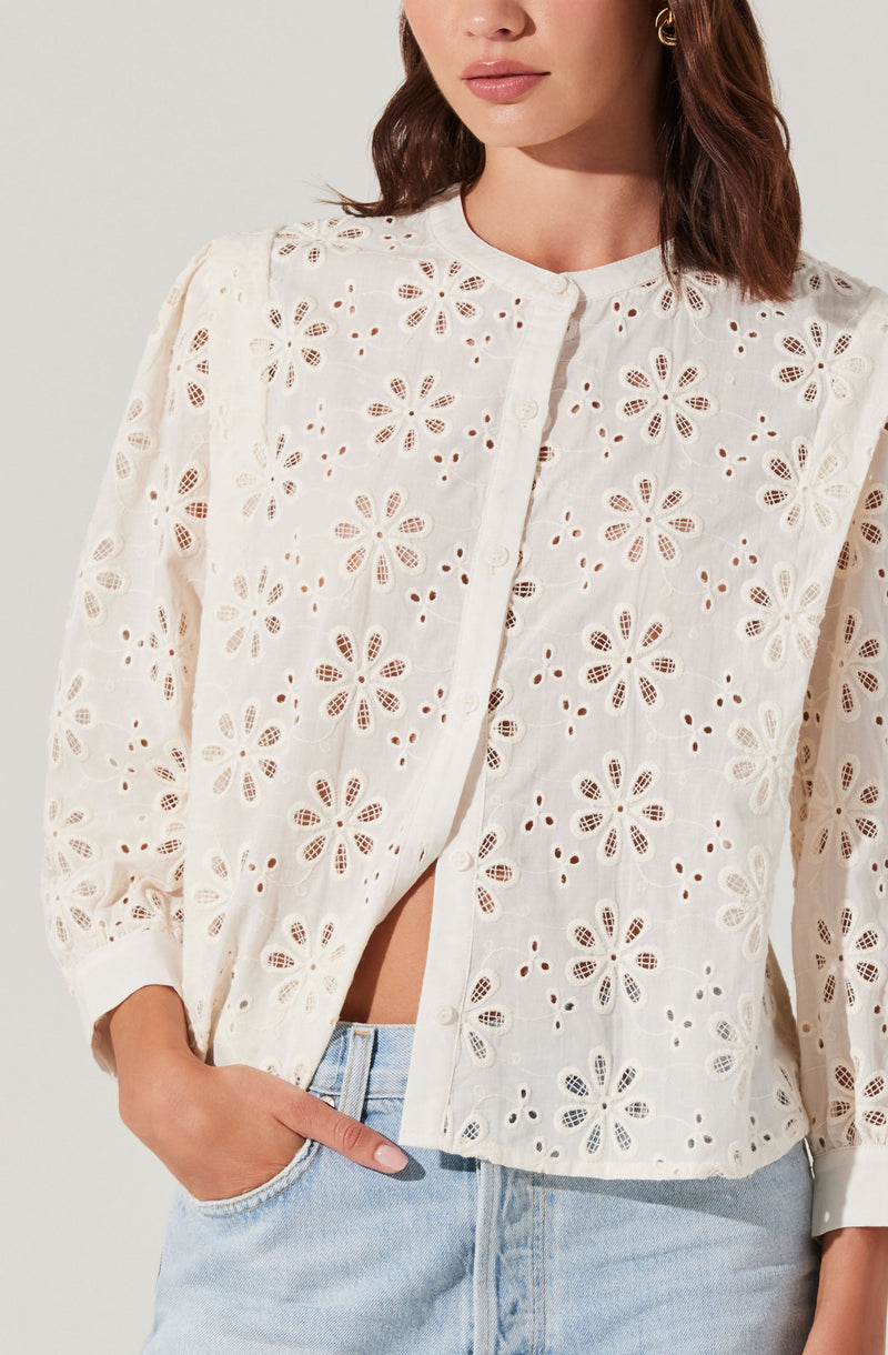 A woman is wearing a white, long-sleeved, button-up blouse made of fabric with an embroidered floral eyelet pattern. The blouse features a round neckline and is slightly open at the bottom, revealing a glimpse of her midriff. She is also wearing light blue jeans.