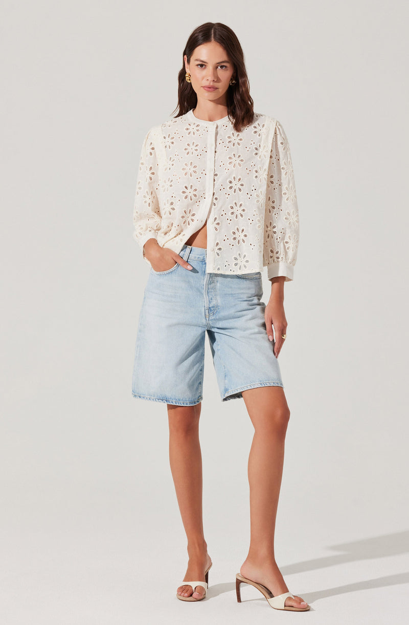 A woman is wearing a white, long-sleeved, eyelet lace jacket with a stand collar and button-up front. The jacket has a loose fit, slightly puffed shoulders, and the sleeves are gathered at the cuffs. She pairs it with light blue, knee-length denim shorts and beige open-toe high-heeled sandals.