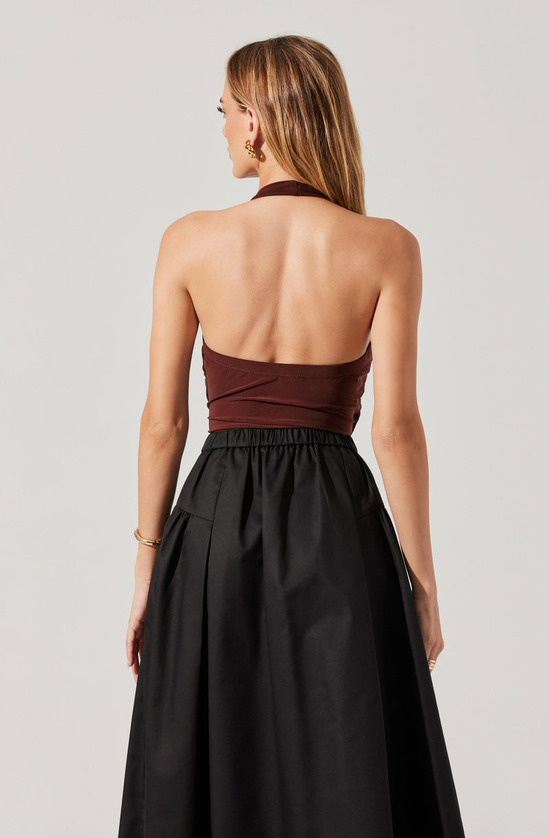 A woman is wearing a sleeveless halter top in dark brown with a gathered detail around the waist, paired with a high-waisted, full-length black skirt that has an elastic waistband and subtle pleats. She has long, straight hair and is accessorized with a gold earring and bracelet.