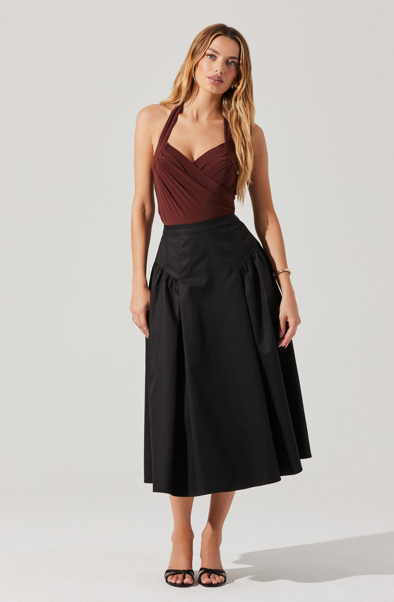 A woman is wearing a sleeveless, deep red halter top with gathered details on the front, paired with a high-waisted, black midi skirt featuring gathered fabric at the hips and a slight flare at the hem. She is also wearing black open-toe high-heeled sandals and a bracelet on her right wrist.