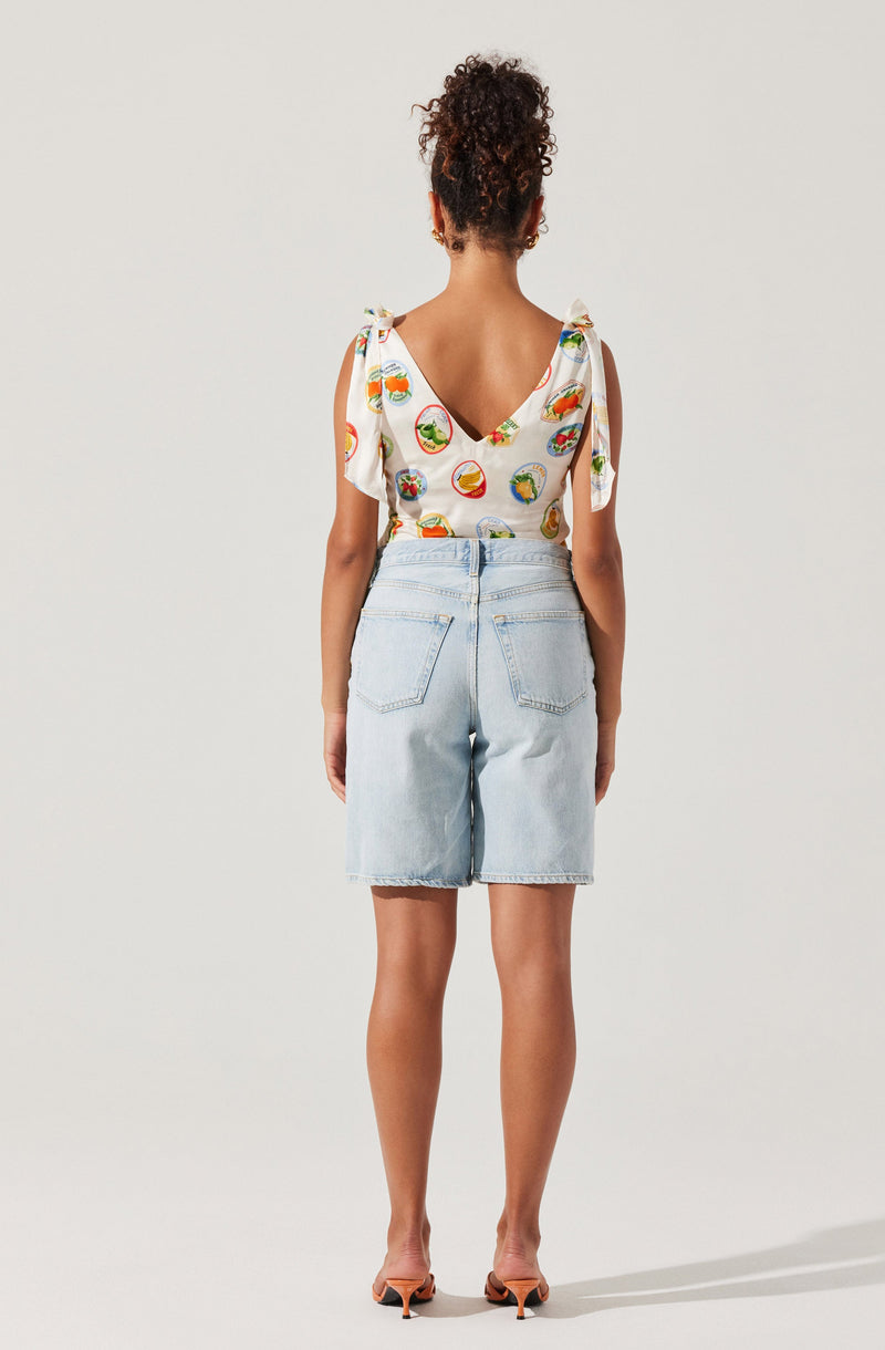 A woman is wearing a sleeveless top with shoulder ties, featuring a colorful pattern of various fruit illustrations, and light-wash denim shorts with back pockets. She is standing with her back to the camera against a plain, light-colored background. She is also wearing open-toe sandals with a small heel.