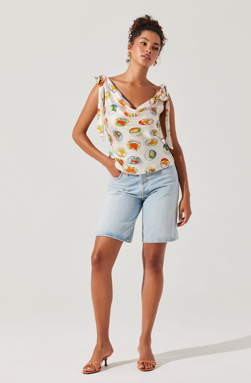 A woman is wearing a sleeveless, cream-colored top with tied shoulder straps and a colorful, circular pattern. She pairs it with light wash denim shorts and tan, open-toe heels.