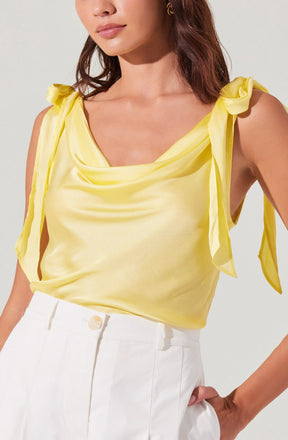 A woman is wearing a sleeveless yellow satin top with tie shoulder straps that form bows, and high-waisted white trousers with a button closure.