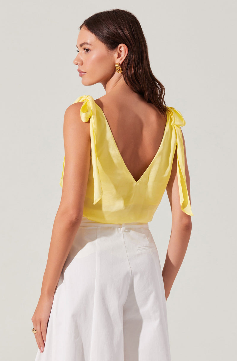 A woman is wearing a sleeveless, yellow top with shoulder ties that form bows, and white high-waisted, wide-leg pants. The top has a deep V-back, and she is accessorized with gold hoop earrings.