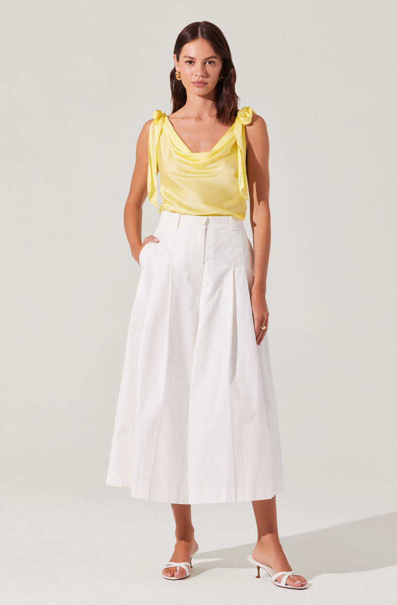 A woman is wearing a sleeveless yellow top with shoulder ties and a draped neckline. She is paired with high-waisted, wide-leg white pants that have pleats and large front pockets. She completes the outfit with white heeled sandals with thin straps.