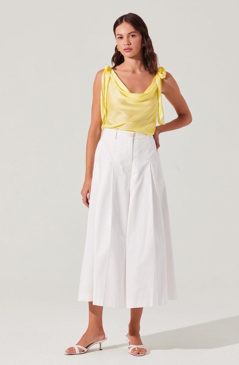 A woman is wearing a sleeveless yellow satin top with shoulder ties and a draped neckline. She pairs it with high-waisted, wide-leg white culotte pants that feature front pleats. She completes the look with white open-toe high-heeled sandals.