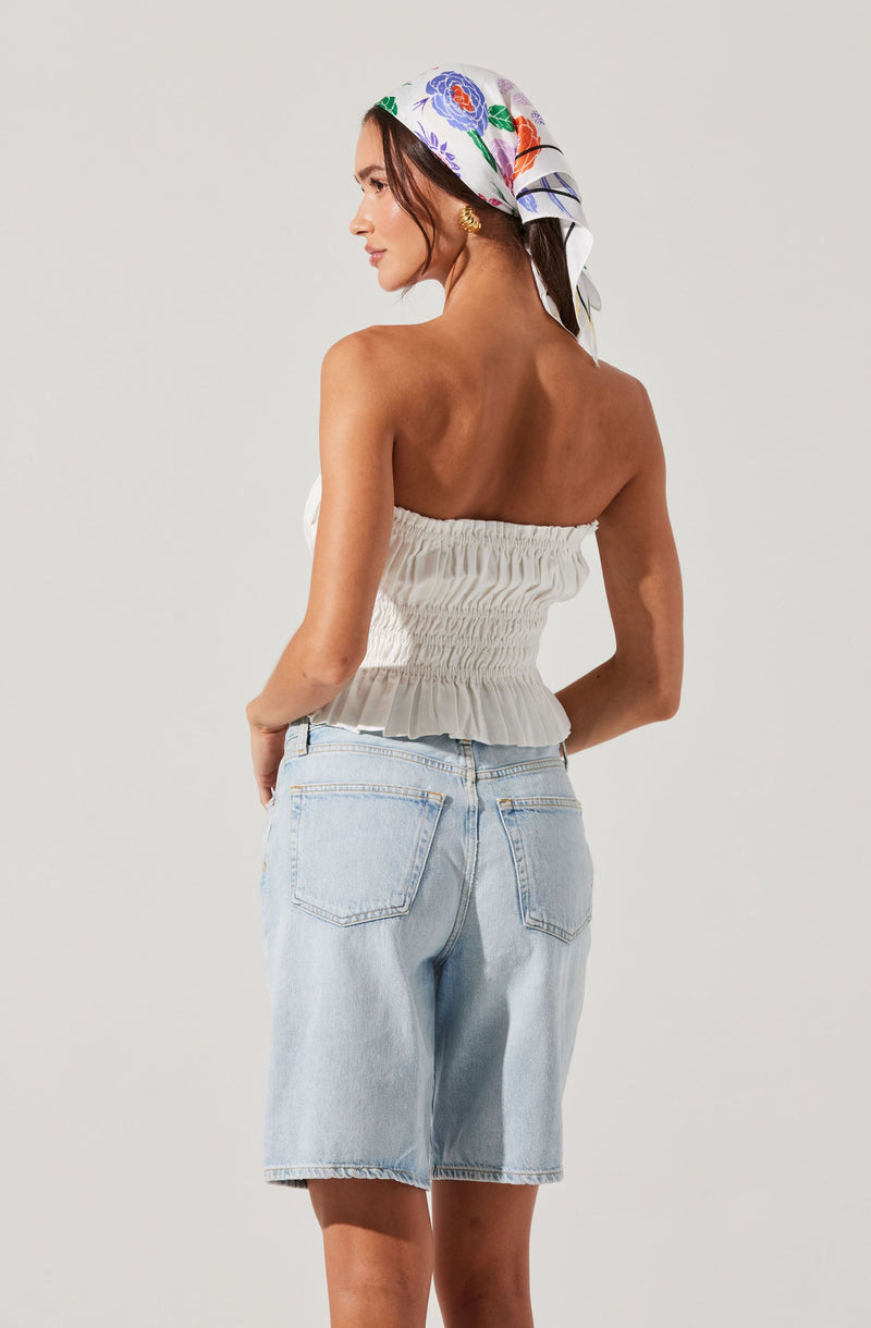 A woman is wearing a white, strapless, ruched top with an elasticized, smocked design at the waist. She is also wearing light-wash denim shorts with a relaxed fit and rolled-up hems. Additionally, she has a colorful, floral-patterned headscarf tied around her head.