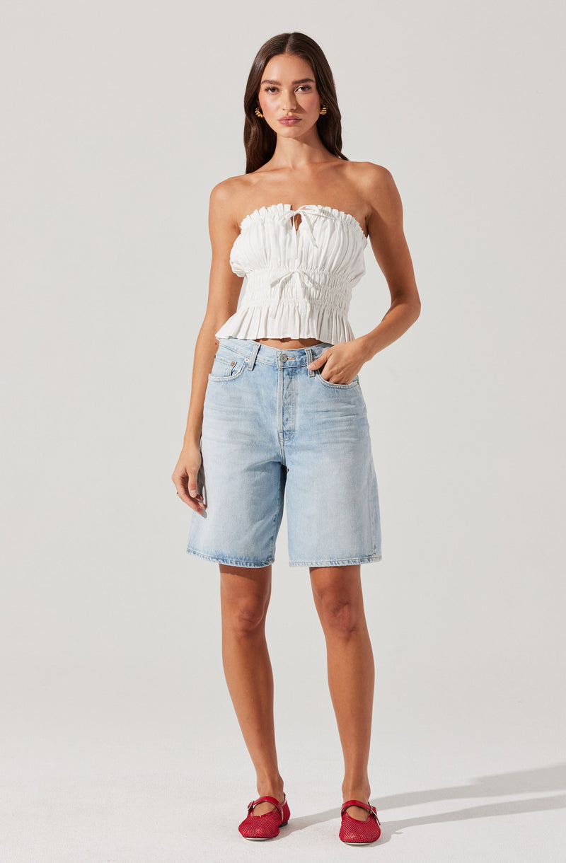 A woman is wearing a strapless, white, ruffled crop top with a gathered, elasticized waist and a small keyhole opening at the front. She is also dressed in light blue, knee-length denim shorts with a classic five-pocket design. She is wearing red flat shoes with a bow detail.