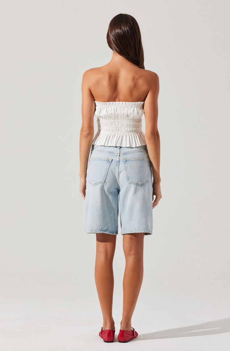 A woman is wearing a strapless, white, ruched top that is fitted and ends above the waist. She is also wearing light blue, knee-length denim shorts with visible stitching and back pockets. Her shoes are red, slip-on style, with a small bow detail on the top. She has long, straight brown hair.