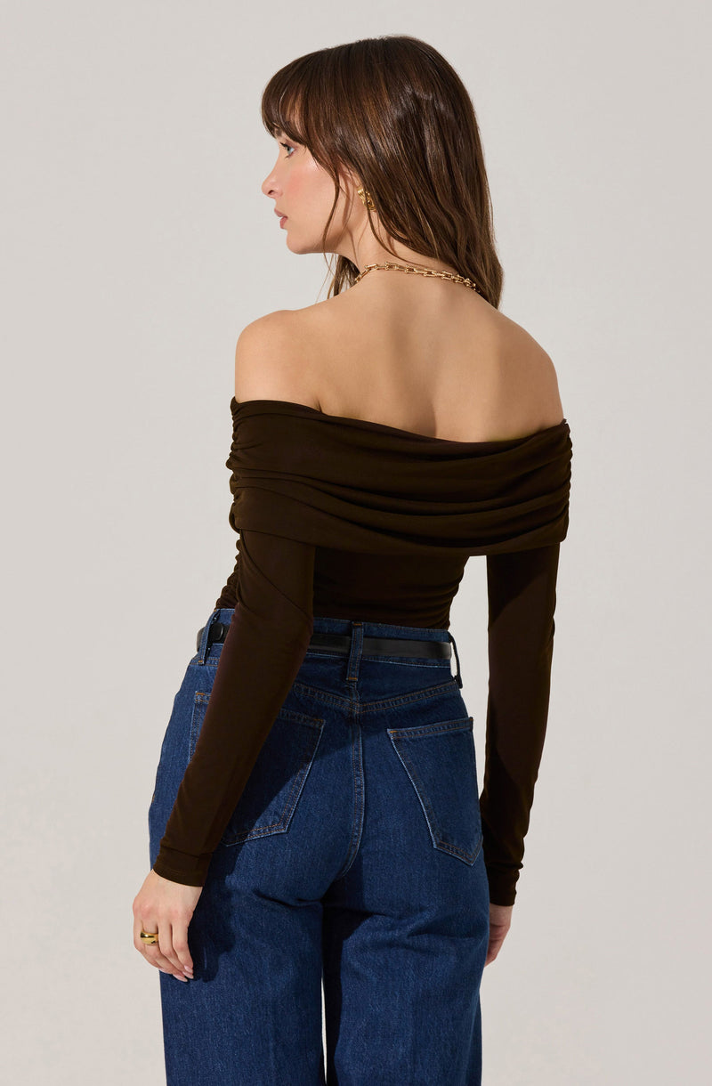 A woman is wearing an off-the-shoulder black top with gathered fabric around the neckline and sleeves. She has paired the top with high-waisted blue jeans. She is accessorized with a gold chain necklace and a ring on her left hand.