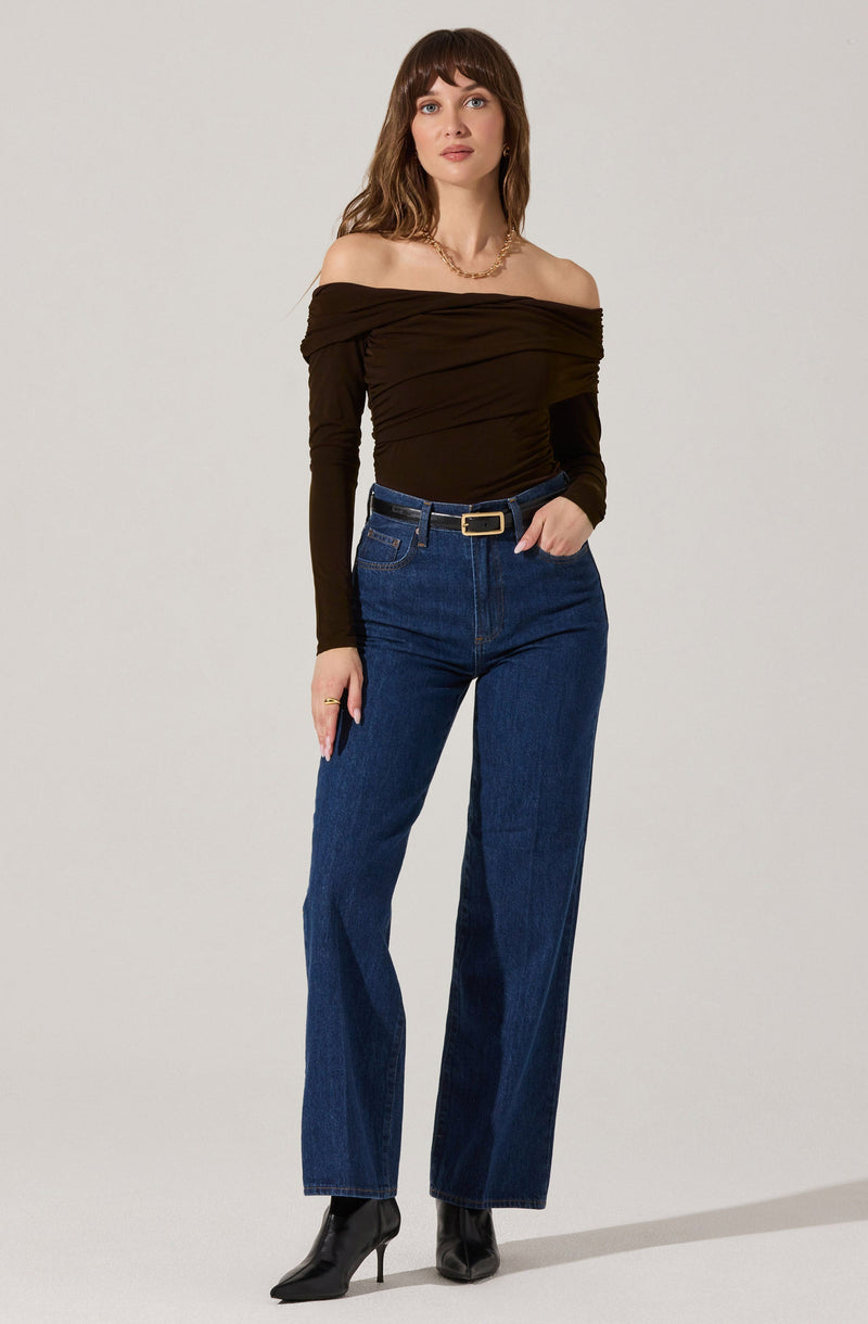 A woman is wearing a dark brown, off-the-shoulder top with ruched detailing. She has paired it with high-waisted, straight-leg blue jeans with a black belt that has a rectangular gold buckle. She completes the look with black heeled boots.