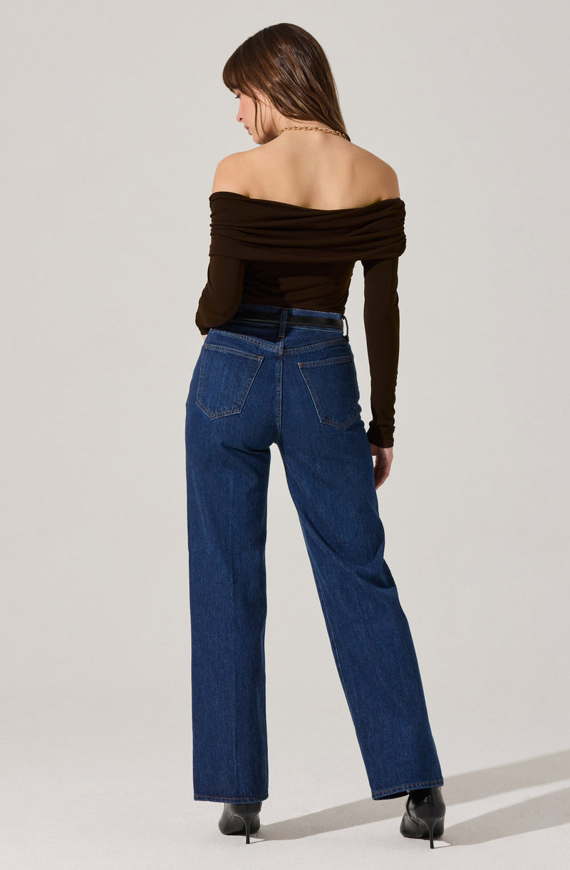 A woman is wearing a black off-the-shoulder top with long sleeves, featuring gathered fabric detail across the shoulders and upper arms. She pairs it with high-waisted, loose-fitting blue jeans and black heeled shoes.