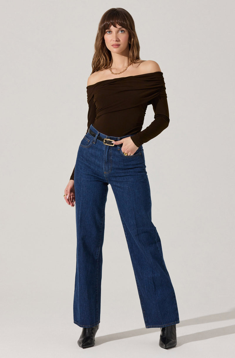 A woman is wearing an off-the-shoulder black long-sleeve top with ruched detailing, paired with high-waisted blue straight-leg jeans and black ankle boots. She has a gold necklace and a gold ring, and her hair is styled with bangs and loose waves.
