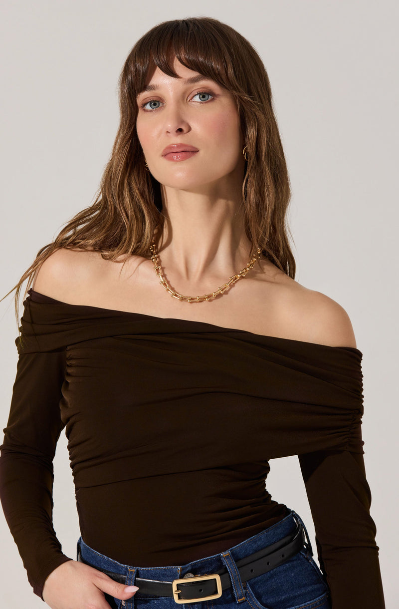 A woman is wearing a dark brown, off-the-shoulder, long-sleeve top with ruched details across the chest. She has paired it with high-waisted blue jeans and a black belt with a gold buckle. She is also wearing a gold chain necklace and small gold hoop earrings.