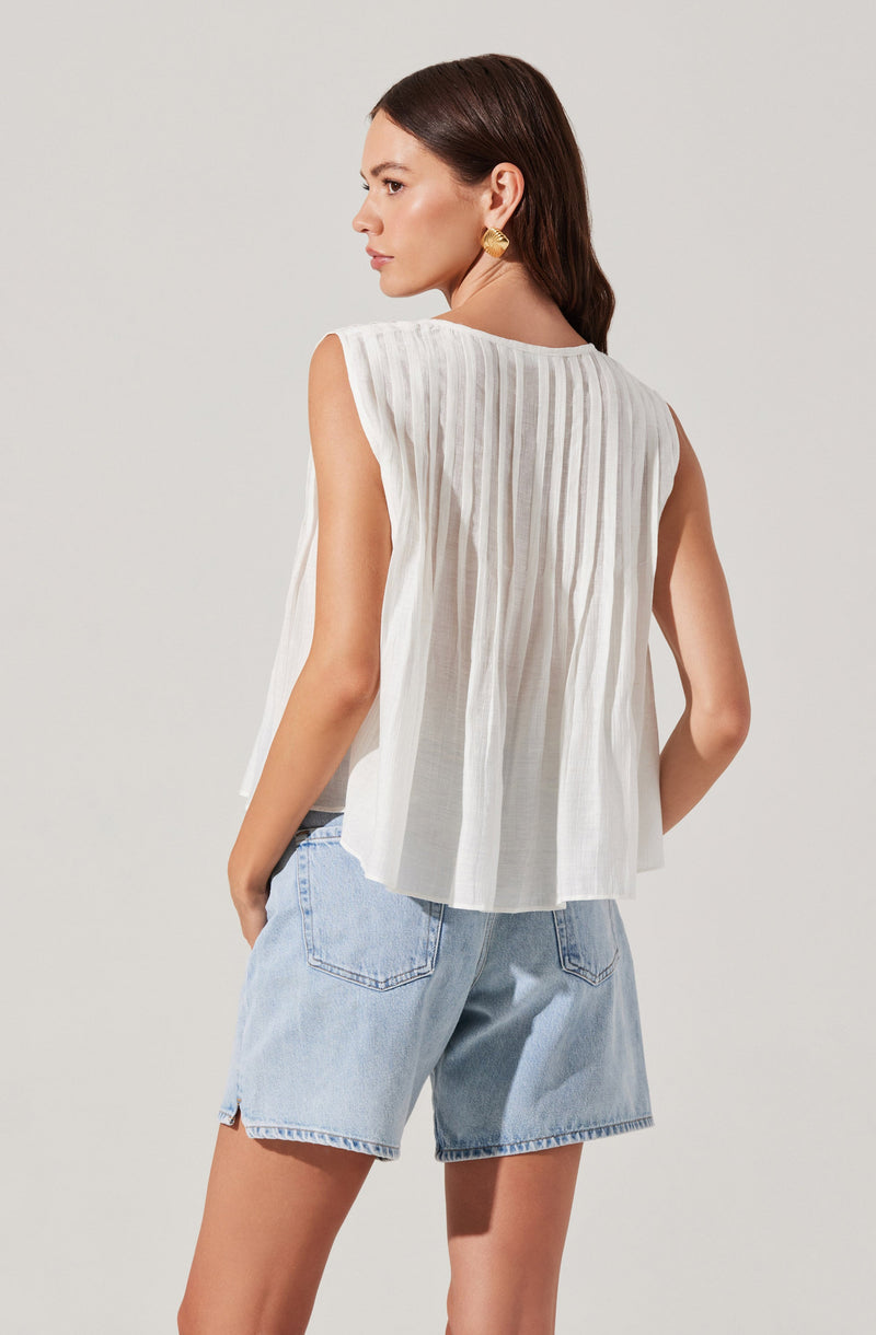 A woman is wearing a sleeveless white top with vertical pleats on the back and light blue denim shorts. She has long brown hair, gold earrings, and is standing against a plain light background.