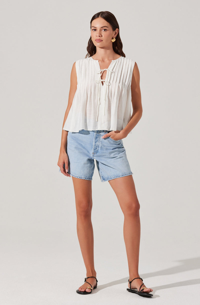 A woman is wearing a sleeveless white top with vertical pleats and a front tie detail, paired with light-wash denim shorts. She is also wearing black strappy flat sandals and has gold hoop earrings.