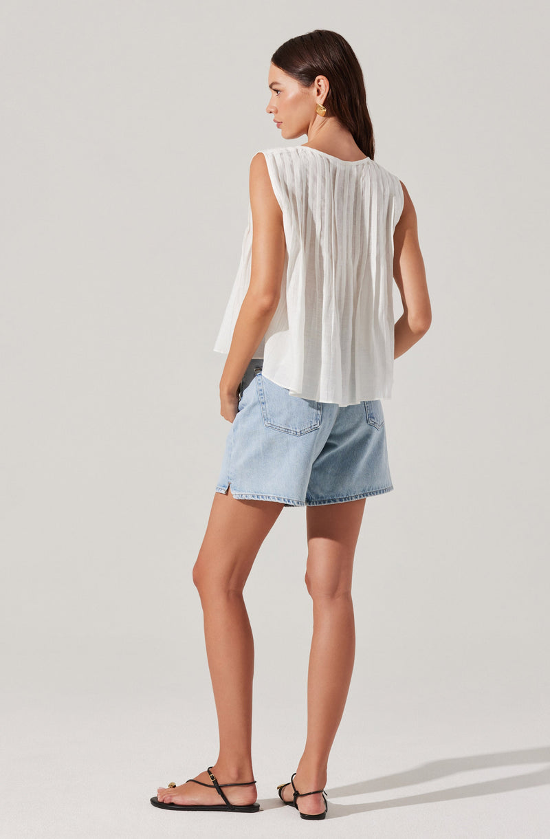 A woman is wearing a sleeveless, white, pleated top with a loose fit and a round neckline. She pairs it with light blue denim shorts that have a relaxed fit and rolled hems. She completes the outfit with black flat sandals that have thin straps.
