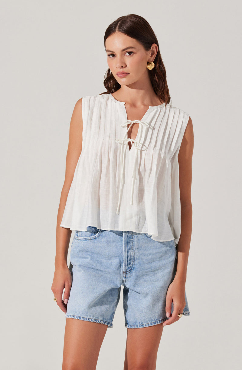 A woman is wearing a sleeveless, cream-colored, lightweight top with vertical pleats and a front tie detail at the neckline. She pairs it with light blue denim shorts.