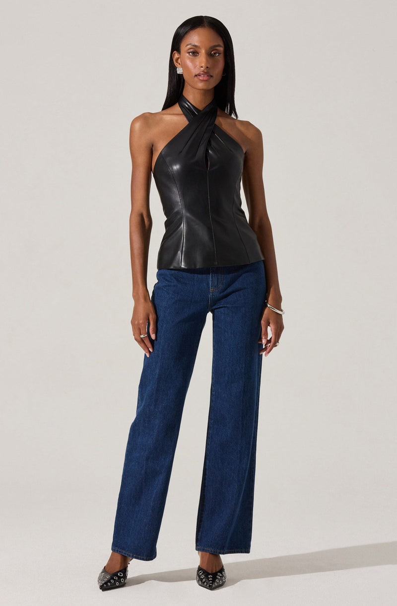 A woman is wearing a sleeveless black leather top with a halter neckline that twists into a knot at the neck, paired with high-waisted, straight-leg blue jeans. She is also wearing black pointed-toe heels with a studded pattern, and accessorized with small earrings and a bracelet.