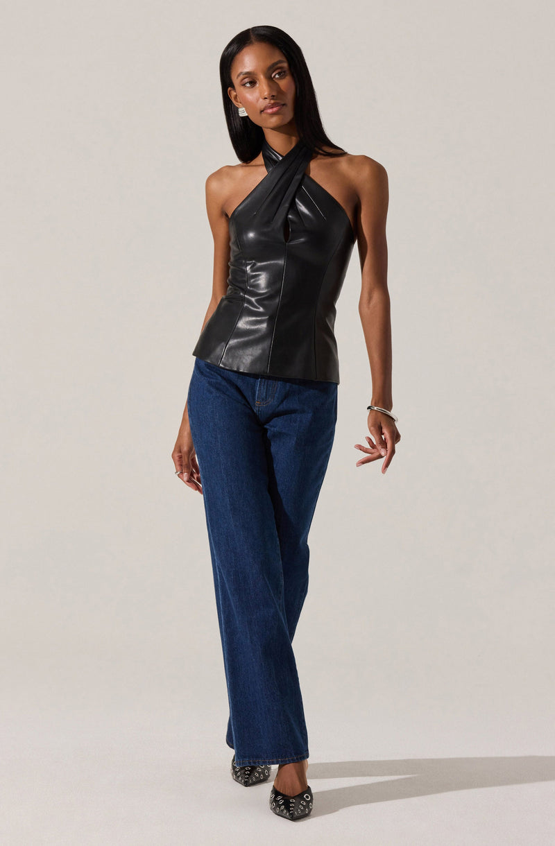 A woman is wearing a black, sleeveless, leather top with a halter neck style that crosses over at the front. She pairs it with high-waisted, straight-leg blue jeans. She completes her outfit with black heels featuring decorative embellishments.
