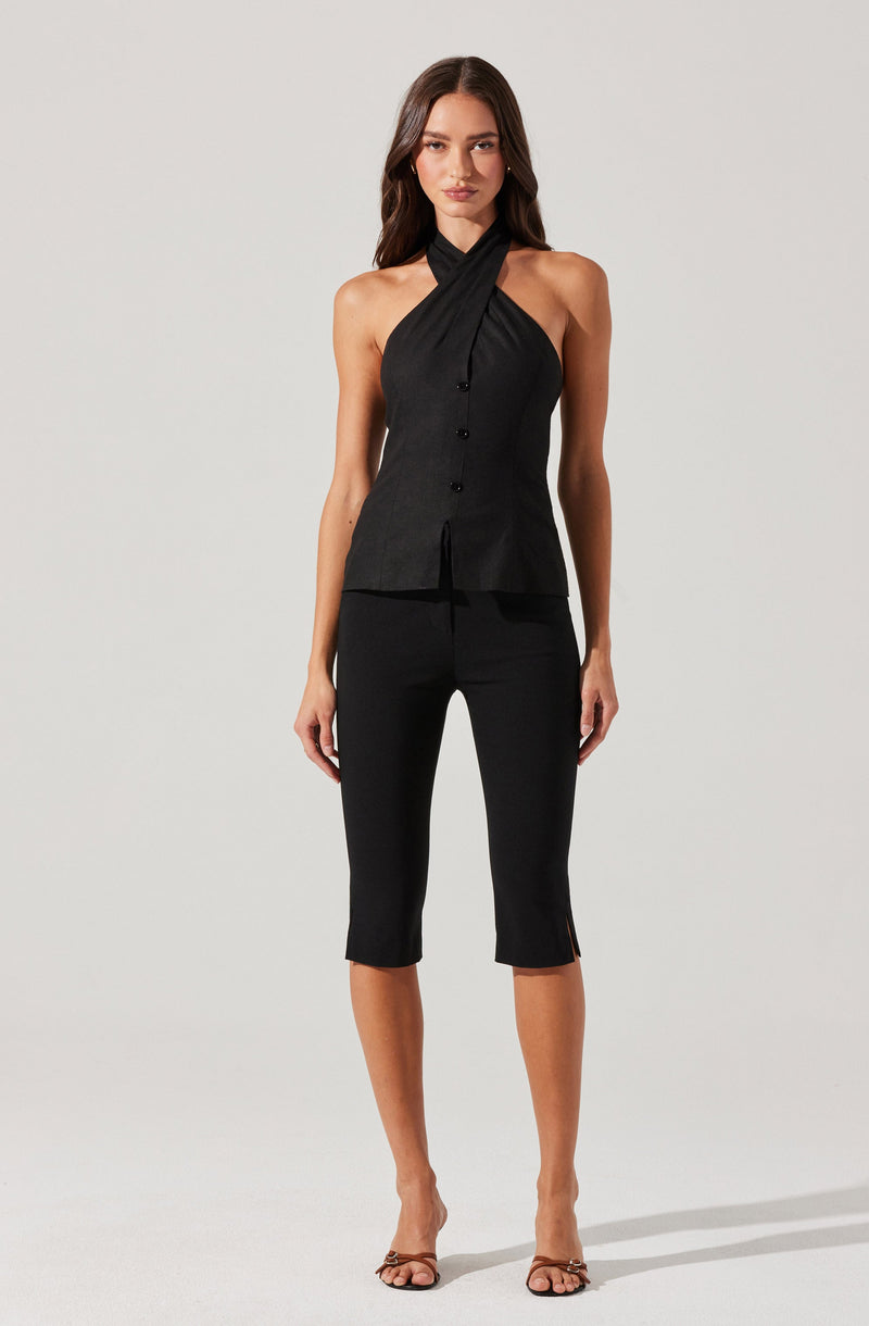 A woman is wearing a sleeveless black top with a high neckline that crosses over at the front, secured with buttons down the center. She pairs it with black cropped pants that have small slits at the bottom hems. The outfit is completed with open-toed high-heeled sandals.