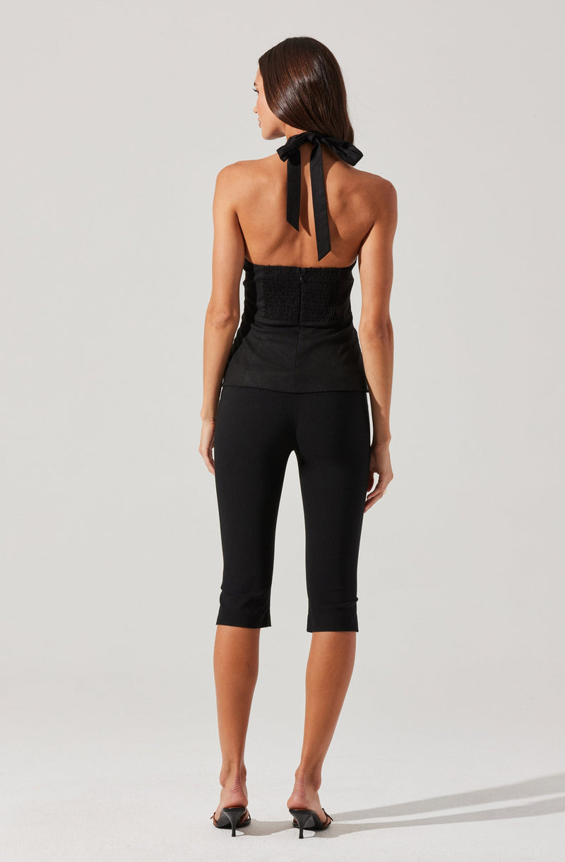 A woman is wearing a sleeveless black top with a textured front and a halter neckline tied with a large bow at the back of her neck. She pairs it with black cropped pants that fit snugly and have a slightly tapered hem, ending below the knees. She is also wearing black high-heeled sandals.