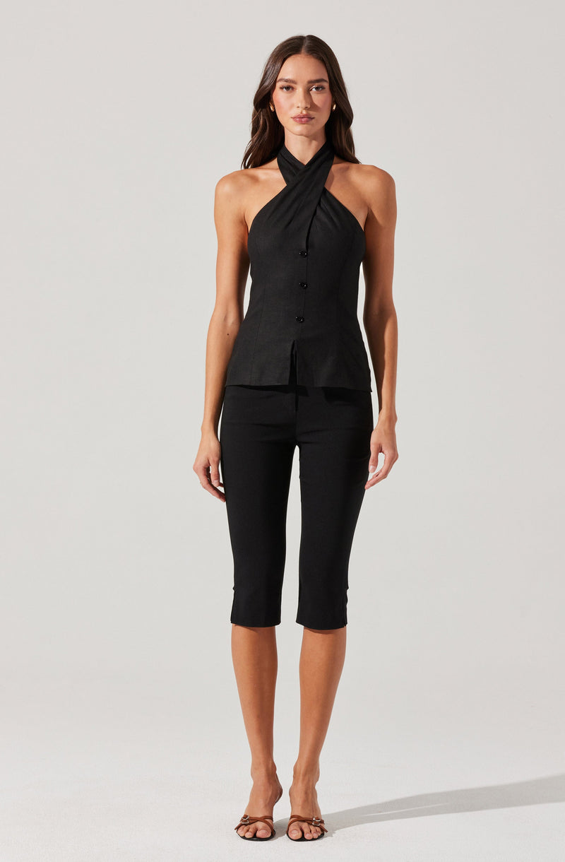 A woman is wearing a sleeveless black top with a halter-neck style that crosses over at the front and buttons down the center, paired with fitted black capri pants. She is also wearing open-toe high heel sandals.
