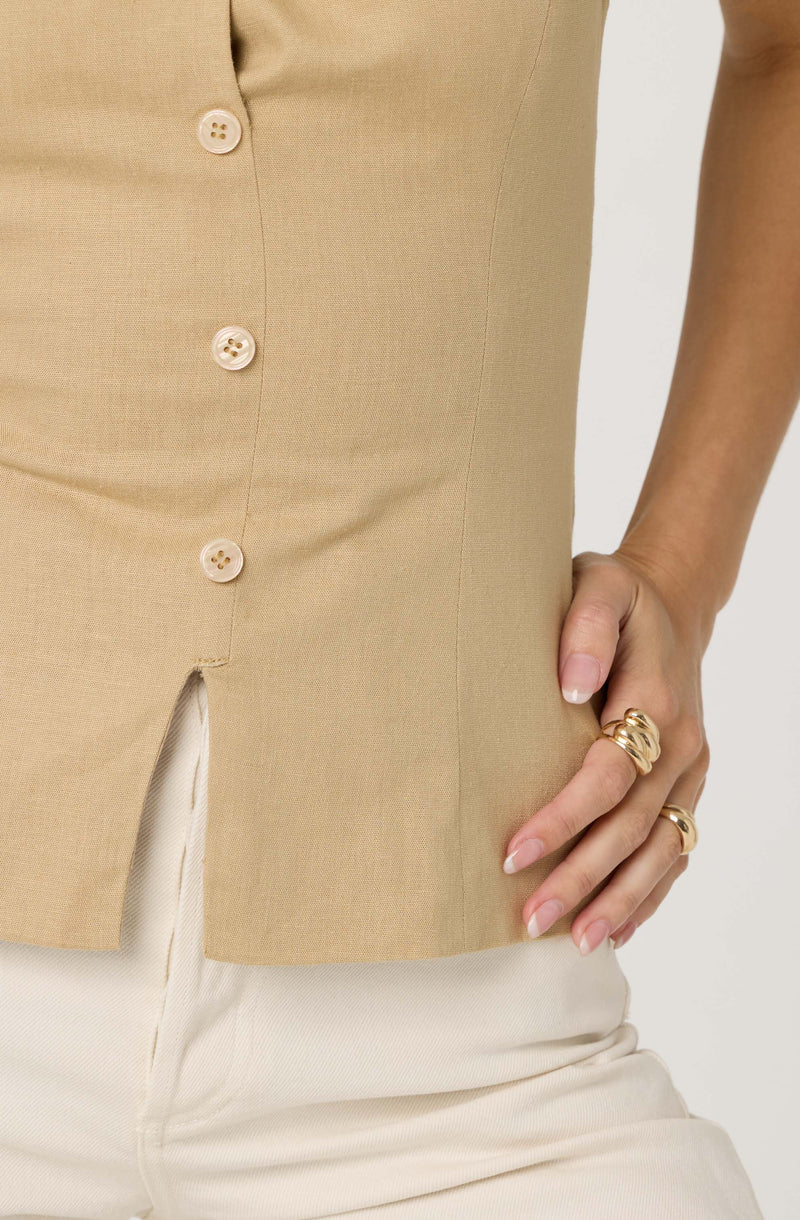 A person is wearing a beige sleeveless button-up top with four light-colored buttons. The top has a small slit at the hem in the front. They are also wearing light-colored pants and have their right hand resting on their hip, adorned with multiple gold rings.