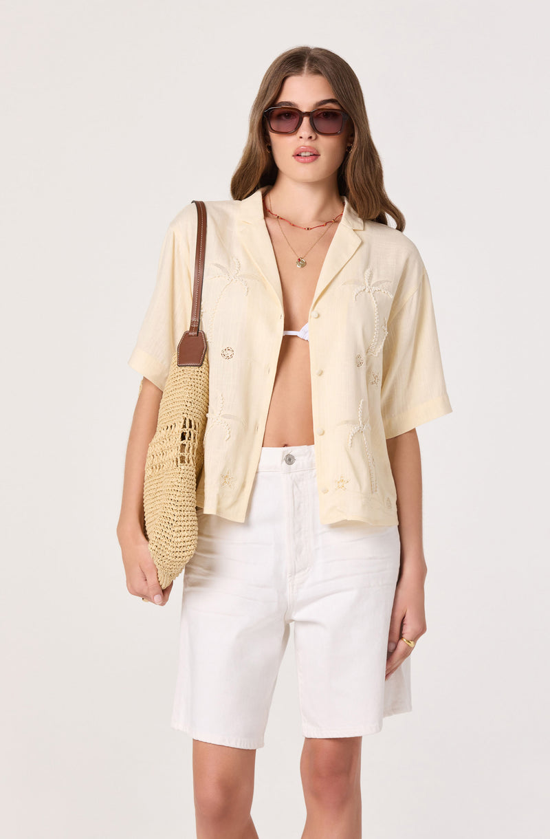 Woman wearing a cream short-sleeve button-up shirt, white shorts, and dark sunglasses, carrying a beige crochet bag.