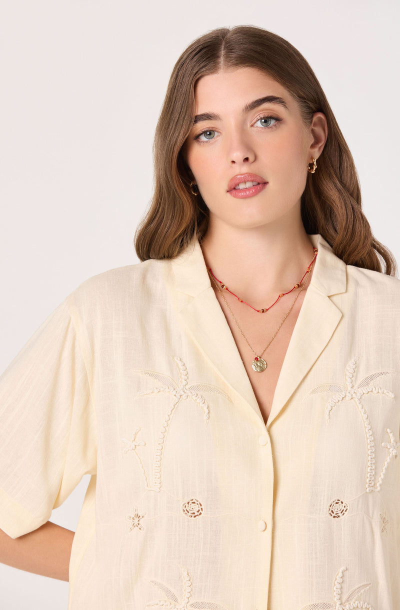 Woman wearing a cream short-sleeve button-up shirt with embroidered palm trees and flowers.