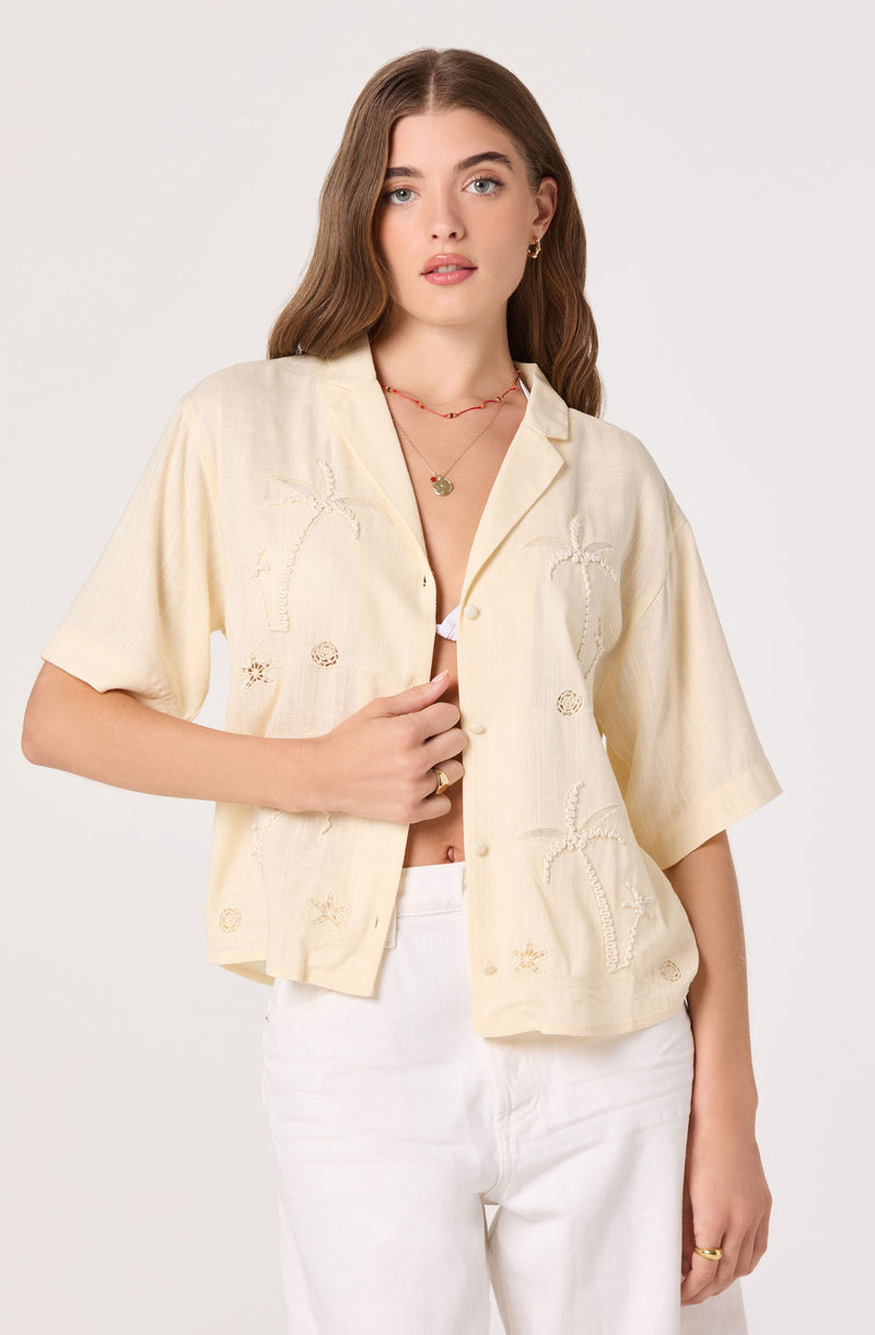 Cream-colored short-sleeve button-up shirt with embroidered palm trees and starfish, worn open over a white top.